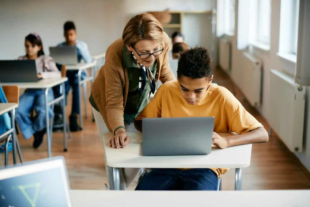 Black elementary student using laptop with help of a teacher in the classroom.