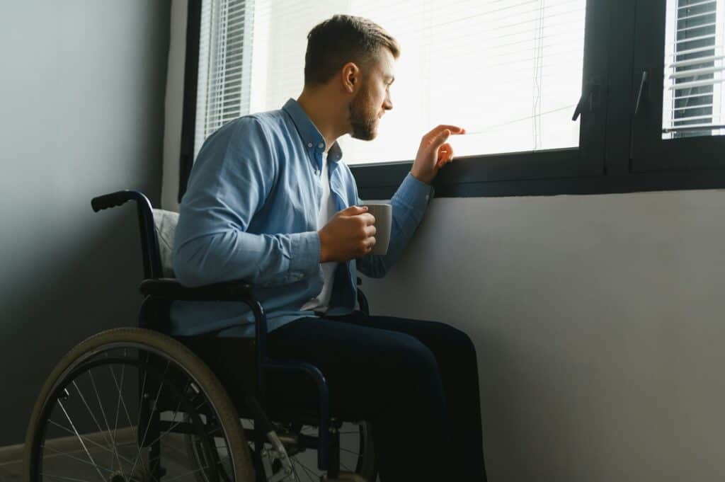 Disabled Person Sits in Wheelchair Against Window. Serious Sad Caucasian Man
