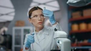 Woman researcher looking drugs in test tube wearing gloves glasses close up.